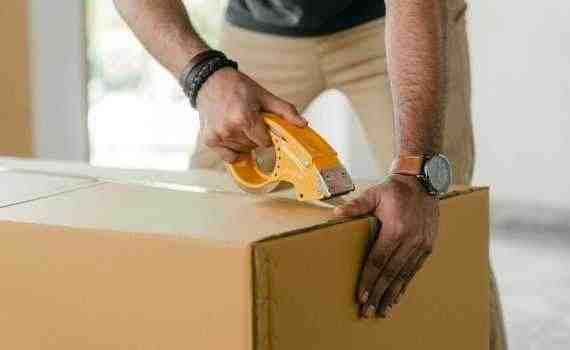 Crop faceless young male with wristwatch using adhesive tape while preparing cardboard box for transportation