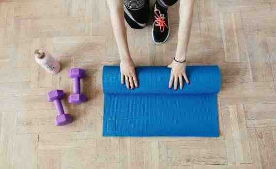Top view of anonymous female athlete in black leggings and sneakers unfolding blue mat for exercising on floor near dumbbells and water bottle in modern fitness center