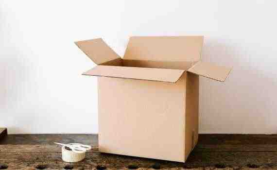 Opened cardboard container placed on old wooden desk near tape and scissors before relocation against white wall