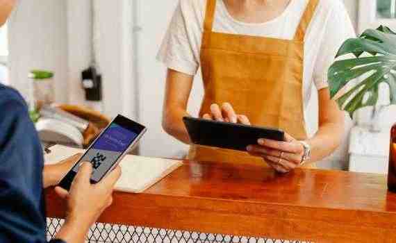 Crop anonymous female employee with application on cellphone screen interacting with partner using tablet at counter in cafeteria