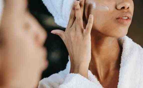 Crop unrecognizable young ethnic female in terry robe applying moisturizing cream on cheek while reflecting in mirror