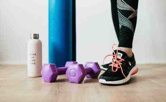 Crop sportswoman wearing sneakers and black leggings standing with sport mat on wooden floor near dumbbells and water bottle before exercising indoors