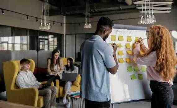 Photo of a Man and a Woman Writing on a Whiteboard with Sticky Notes